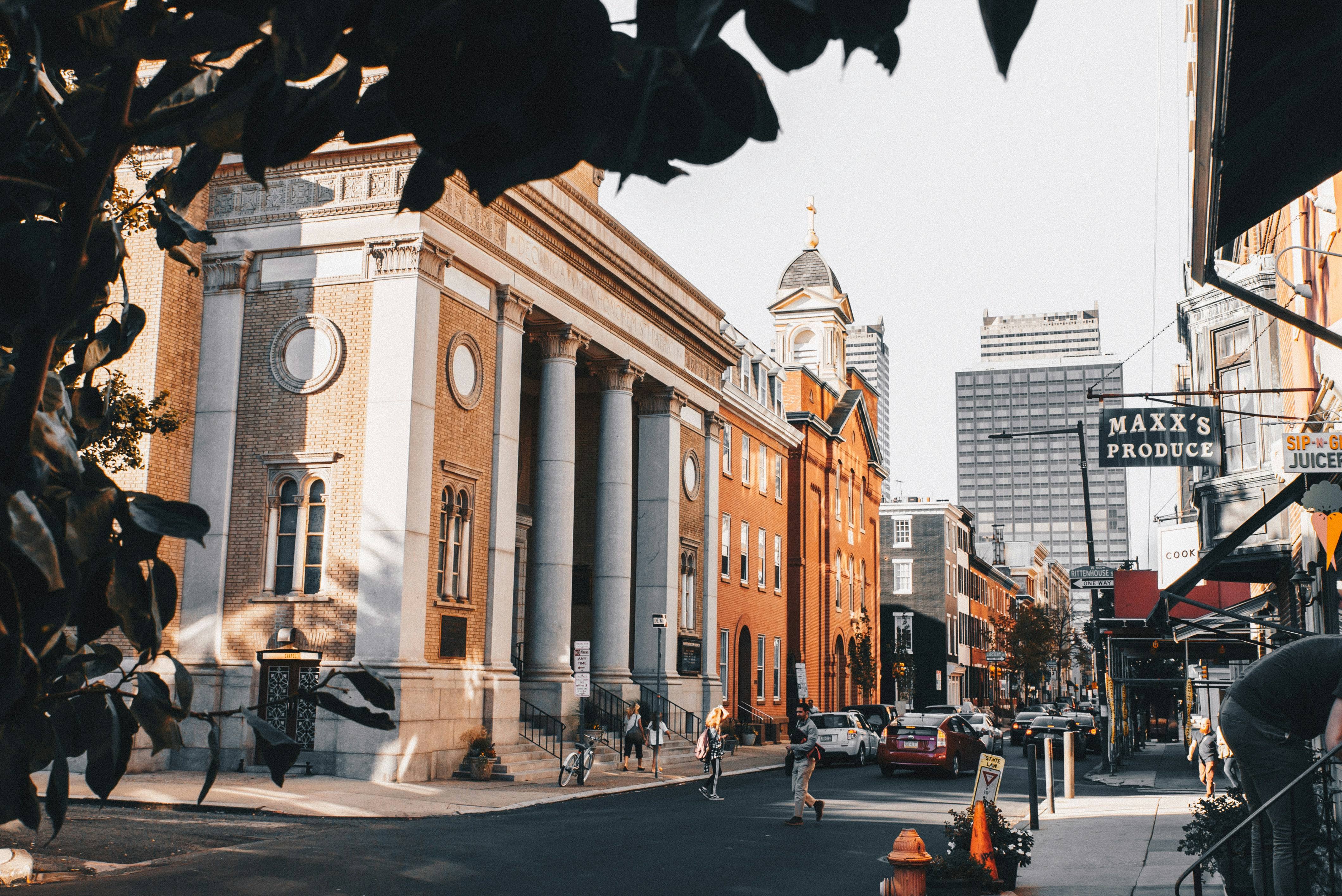 A city street framed by leaves, with a columned stone building and a corner produce market beneath a tall office tower