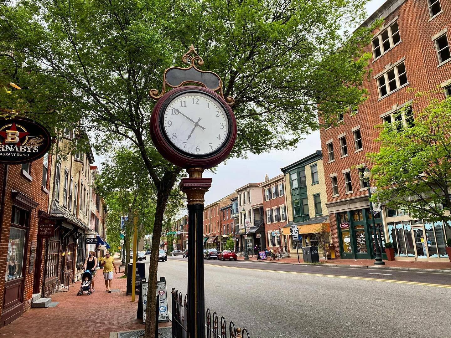 A street clock on the brick sidewalk of Gay Street in downtown West Chester, with shops and restaurants along the block